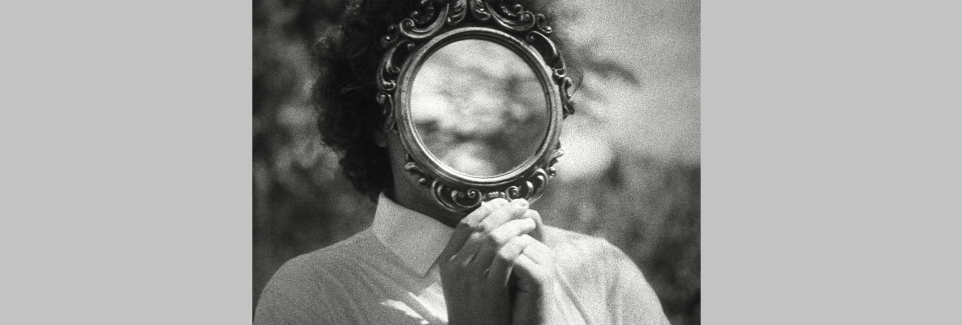 A black and white image of a person in a button-up shirt holding a mirror up so that their face cannot be seen, reflecting instead a part of the sky