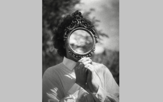 A black and white image of a person in a button-up shirt holding a mirror up so that their face cannot be seen, reflecting instead a part of the sky